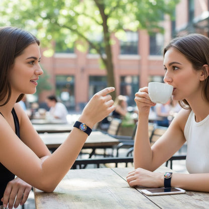 Two women sitting at an outdoor cafe wearing snap button charm anxiety bracelets, one holding a cup and the other gesturing with her hand.